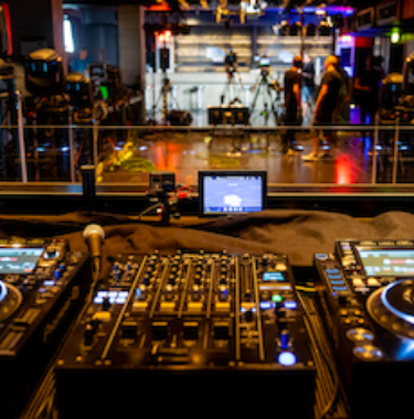 DJ-Pult mit Mischpult und Turntables im Vordergrund, dahinter Kamerateam in Eventlocation mit Bühnenlicht.