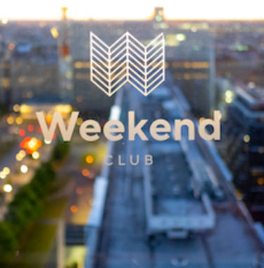 Logo des „Weekend Club“ auf einer Fensterscheibe mit Blick über die Stadt bei Dämmerung.