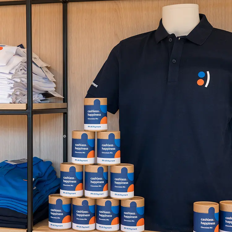 Merchandise-Regal mit gefaltetem VR Payment-Textil, Polohemd auf Büste und gestapelten „cashless happiness“-Dosen.