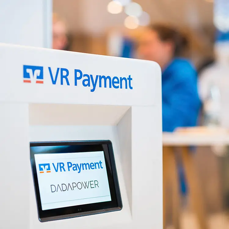 Nahaufnahme eines VR Payment-Terminals mit Displayanzeige „DADAPOWER“. Im Hintergrund unscharf Messebetrieb.