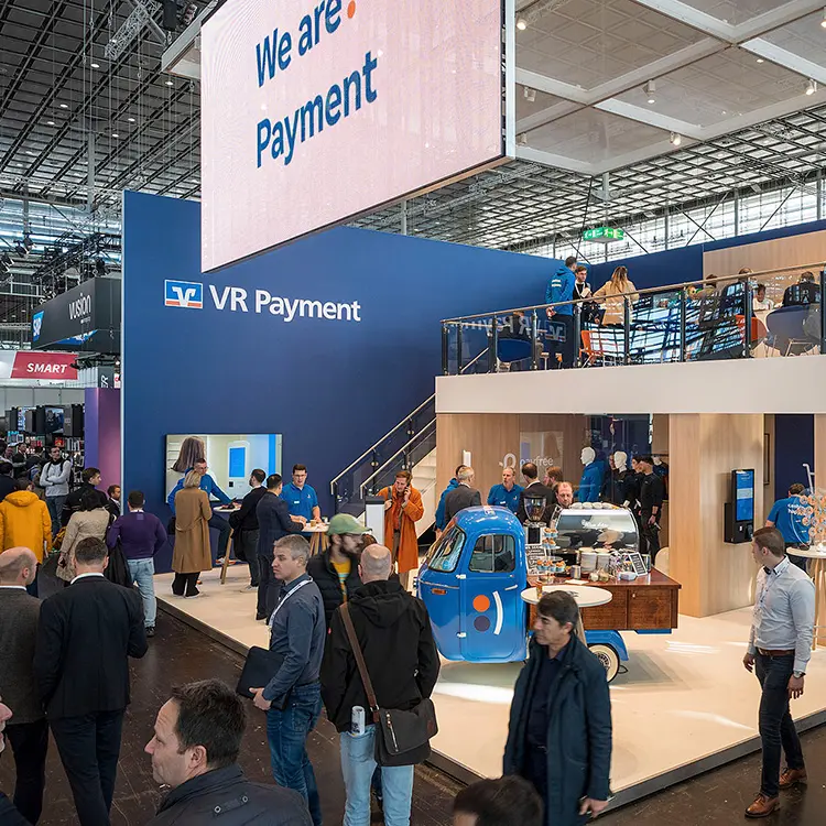 Großer VR Payment-Messestand mit vielen Besucher*innen, digitalem We are Payment-Schild und blauer Kaffeebar im Ape-Fahrzeug.