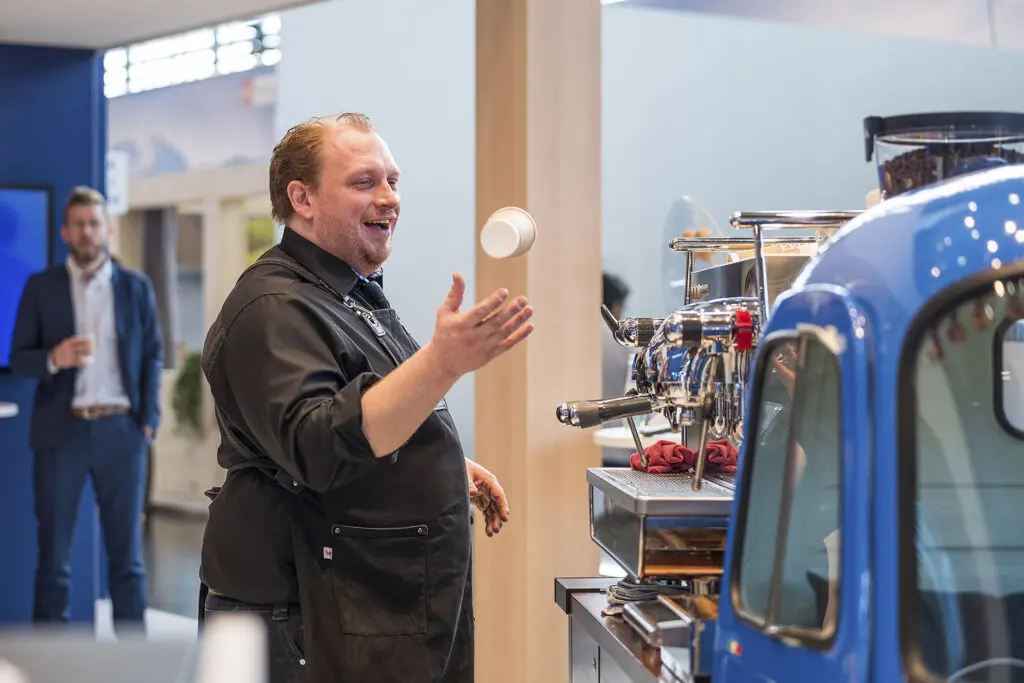 Barista wirft lachend einen Kaffeebecher in die Luft neben einer Siebträgermaschine und einem blauen Verkaufsfahrzeug.