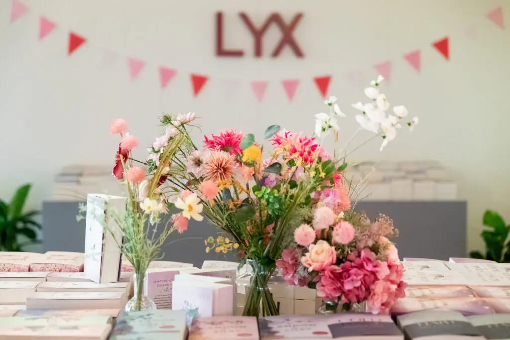 Bücher- und Blumentisch mit LYX-Logo im Hintergrund, dekoriert mit Wimpelgirlande und frischen Blumen.