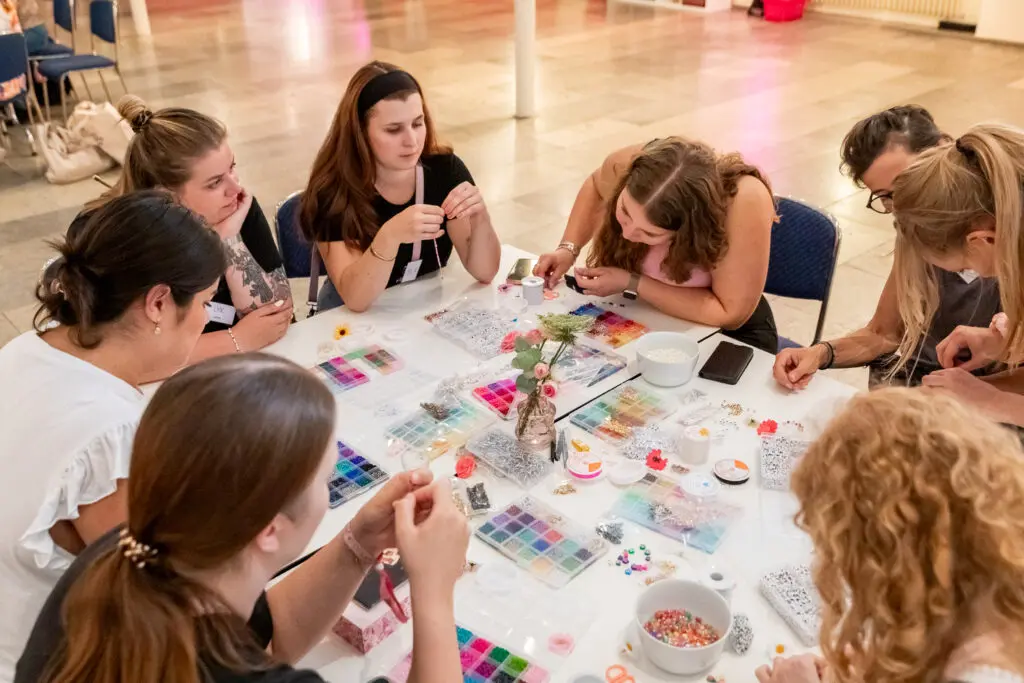 Gruppe junger Frauen sitzt an einem Basteltisch und fädelt bunte Perlen zu Armbändern an der DIY-Armbänder-Station