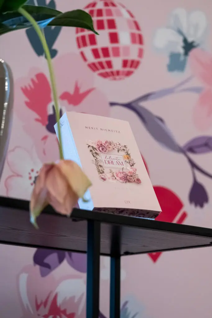 Nahaufnahme eines LYX-Romans von Merit Niemeitz mit floralem Cover, daneben eine herabhängende Blume vor pastellfarbener Wand.