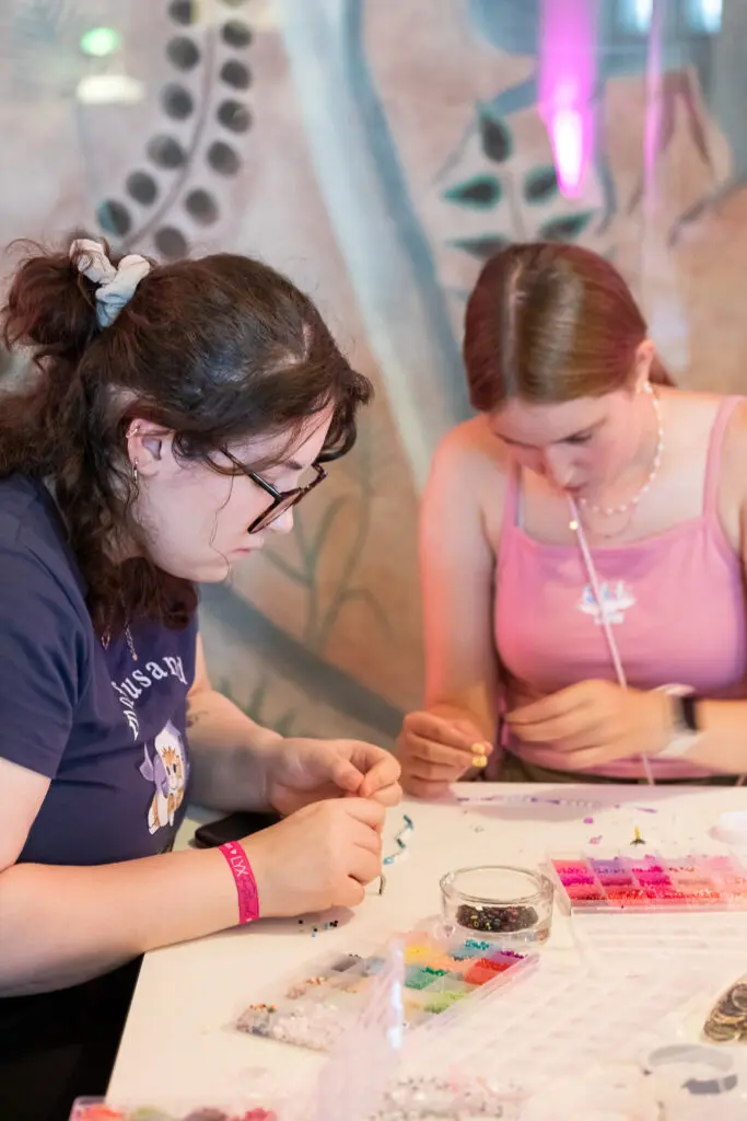 Zwei junge Frauen fädeln konzentriert bunte Perlen an einem Basteltisch mit Sortierboxen und Schalen.