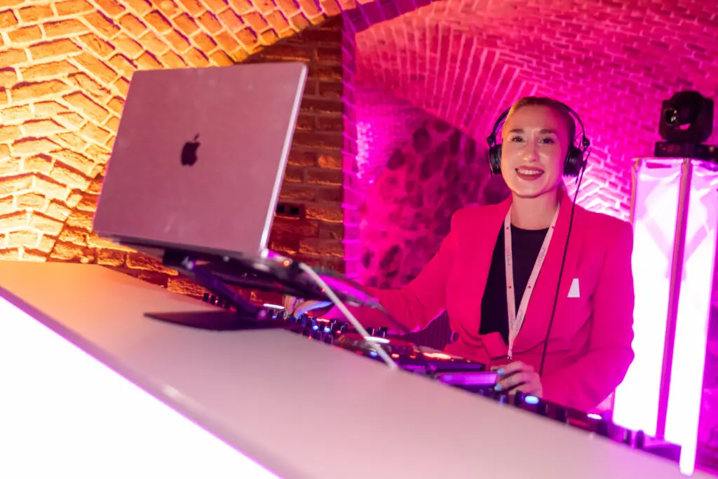DJane in pinkem Blazer steht lächelnd an einem DJ-Pult mit Laptop in einem stimmungsvoll beleuchteten Gewölbekeller.