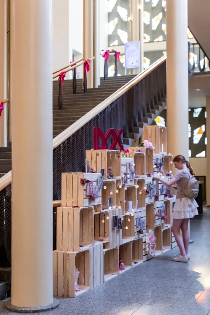 Junges Mädchen betrachtet dekorative Bücherwand aus Holzkisten mit LYX-Logo und Charakterkarten im Treppenhaus.