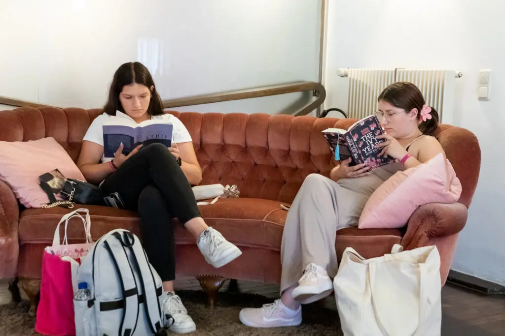 Zwei junge Frauen sitzen entspannt auf einem rosa Sofa und lesen Bücher; um sie herum liegen Taschen mit Festival-Utensilien.