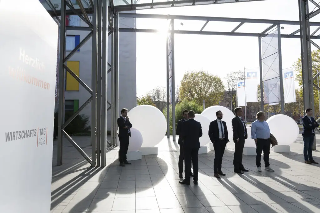Besucher*innen betreten das lichtdurchflutete Foyer eines Kongresszentrums mit Willkommensbeschriftung für den Wirtschaftstag.
