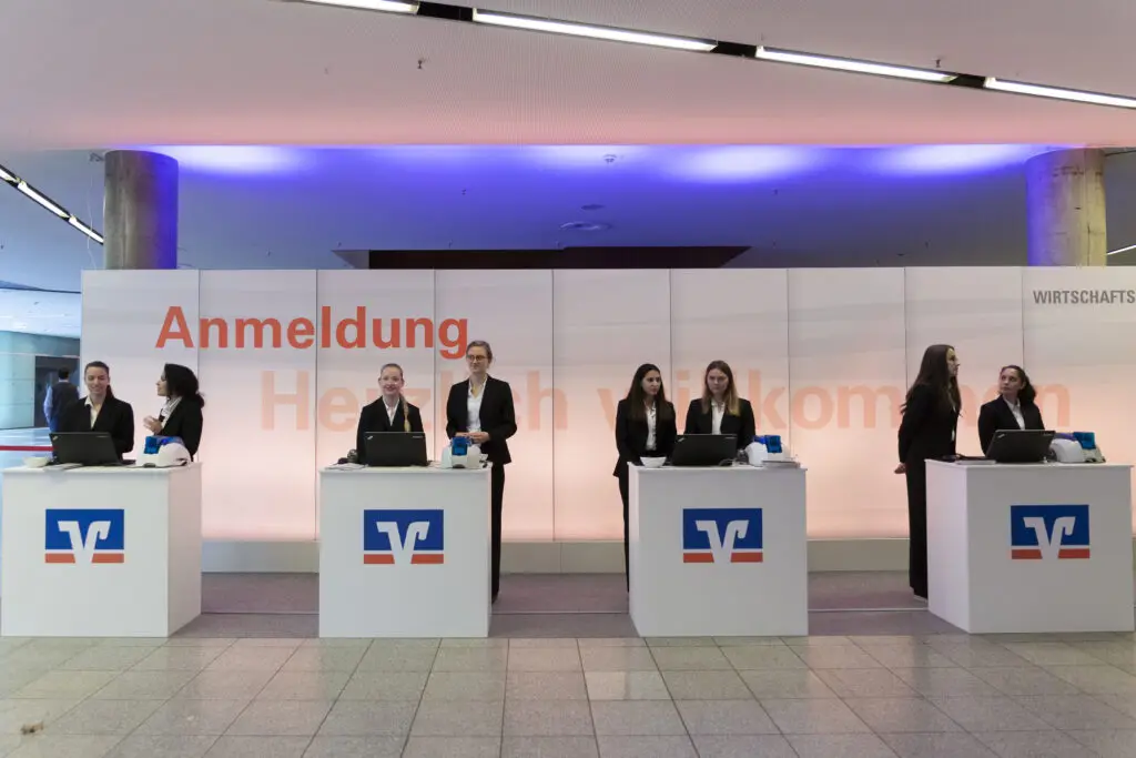 Mehrere Mitarbeiter*innen stehen hinter Anmeldetheken der Volksbanken Raiffeisenbanken und begrüßen Gäste in einem modernen Eingangsbereich.