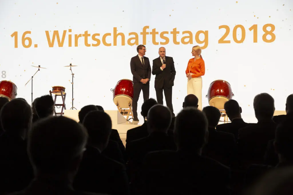 Drei Sprecher stehen auf der Bühne beim 16. Wirtschaftstag 2018 vor einer weißen Leinwand mit goldener Aufschrift.