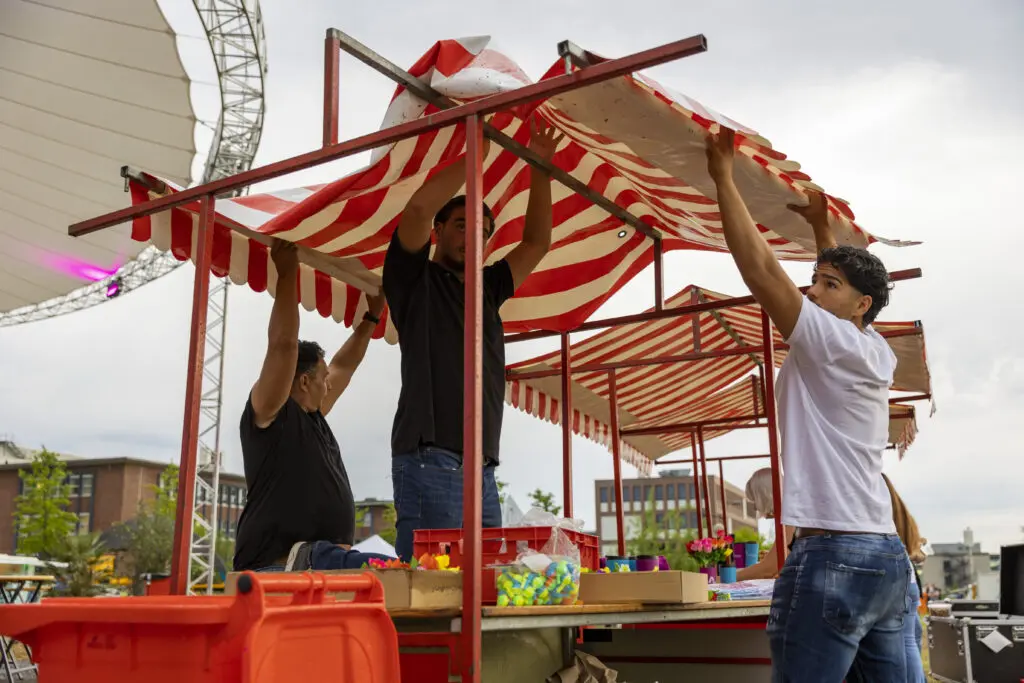 Drei Männer bauen einen rot-weiß gestreiften Marktstand auf, vermutlich für ein Event oder Straßenfest. Auf dem Stand liegen bereits bunte Entchen und Deko – im Hintergrund sind weitere Stände und ein großes Zeltdach zu sehen.