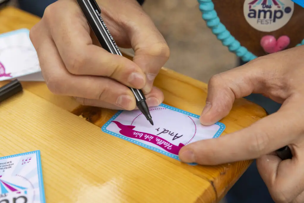 Nahaufnahme von zwei Händen, die ein Namensschild mit der Aufschrift „Hallo, ich bin“ ausfüllen – der Name „Amel P.“ wird gerade geschrieben. Im Hintergrund ist ein Lebkuchenherz mit dem „amp Fest“-Logo sichtbar.