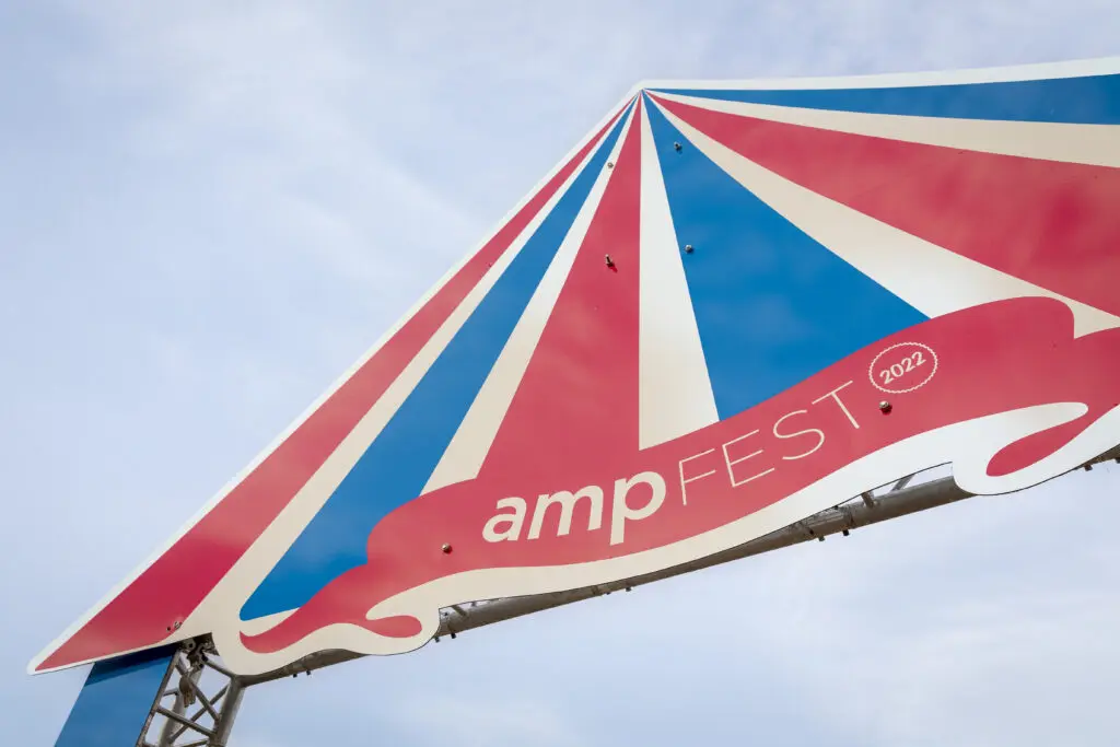 Ein großes, dreieckiges Schild in Zeltform mit rot-blau-weißen Streifen trägt die Aufschrift „ampFEST 2022“ und ist an einer Metallkonstruktion befestigt, vor einem hellblauen Himmel.