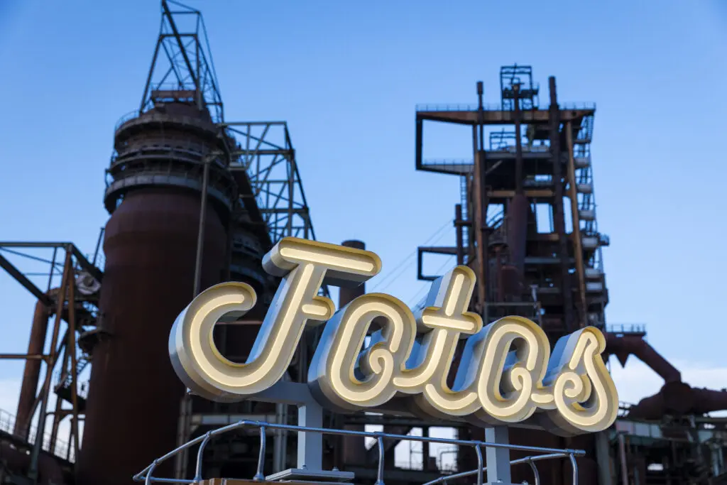 Ein beleuchtetes Schild mit der Aufschrift „Fotos“ in geschwungener Schrift steht im Vordergrund, dahinter ragen rostige Industrieanlagen und ein blauer Himmel empor – eine Kombination aus Retro-Charme und Industrieästhetik.
