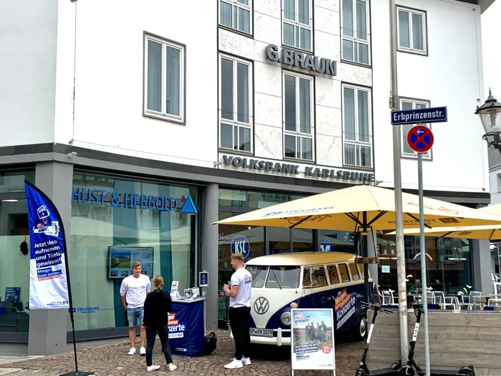 VW Bulli mit Morgen-kann-kommen Volksbanken Raiffeisenbanken Branding und Promotion-Stand mit Liegestühlen und Theke vor einer Bankfiliale