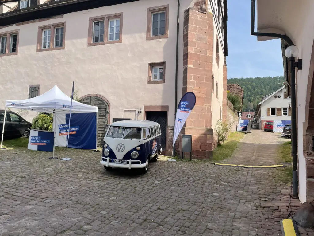 Blauer VW-Bulli mit „Morgen kann kommen“-Branding steht vor historischem Gebäude neben Infozelten und Werbefahnen.