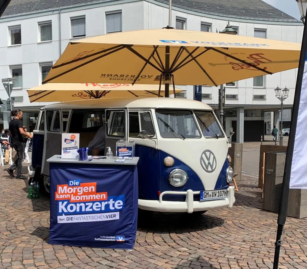 Blauer VW-Bulli mit Infostand und Sonnenschirmen wirbt in der Innenstadt für „Die Morgen kann kommen Konzerte“.
