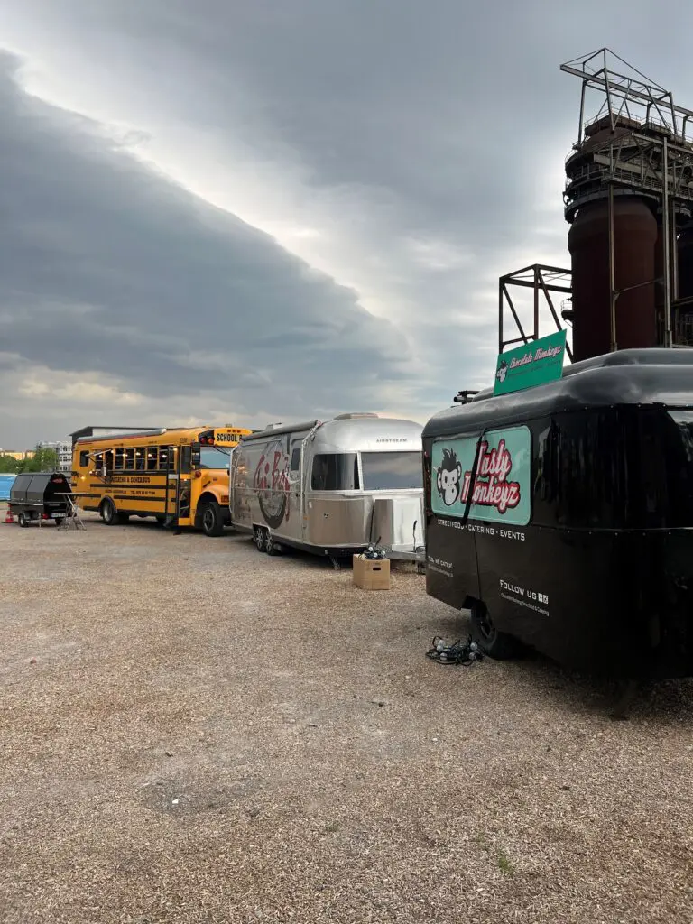 Drei Foodtrucks in einer Reihe auf einem Schotterplatz vor einem alten Industriebau – ein gelber Schulbus, ein silberner Airstream-Anhänger mit „La Pizza“-Schriftzug, und ein schwarzer Anhänger mit dem Logo „Tasty Monkeyz“.