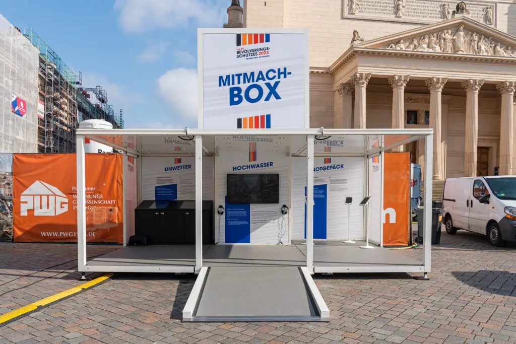 Weiße mobile Ausstellungseinheit mit der Aufschrift „MITMACH-BOX“ des Bevölkerungsschutzes steht auf einem gepflasterten Platz vor historischer Kulisse. Auf den Infowänden sind Themen wie Hochwasser und Stromausfall sichtbar.