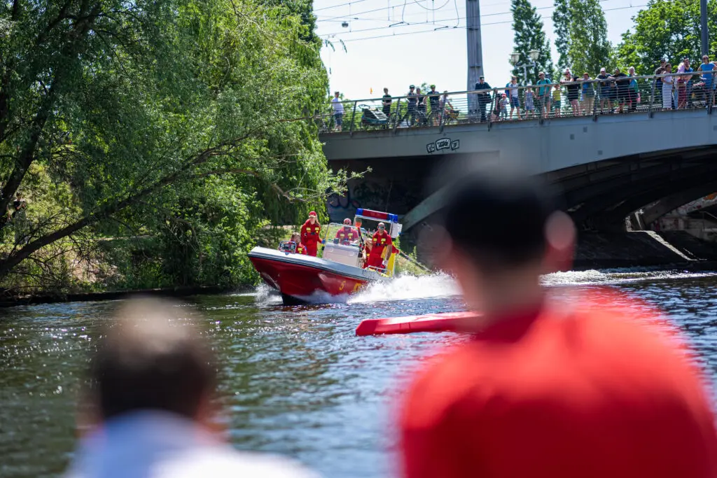 Ein Rettungsboot der Wasserrettung fährt mit hoher Geschwindigkeit über einen Fluss, besetzt mit Einsatzkräften in roter Kleidung. Im Vordergrund beobachten Zuschauer*innen die Szene, während sich auf der Brücke im Hintergrund weitere Besucher*innen versammeln.