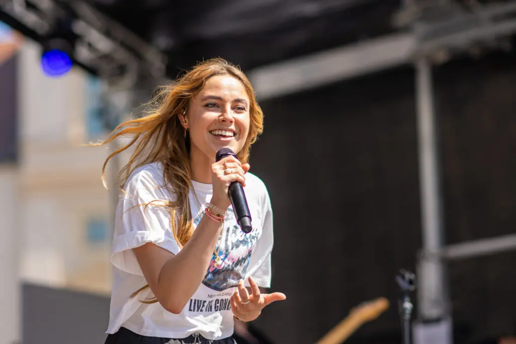 Nahaufnahme der Sängerin "SOPHIA" mit langen, offenen Haaren, die auf einer Bühne in ein Mikrofon singt und dabei lächelt. Sie trägt ein weißes T-Shirt mit Aufdruck, im Hintergrund ist Bühnentechnik leicht unscharf zu erkennen.