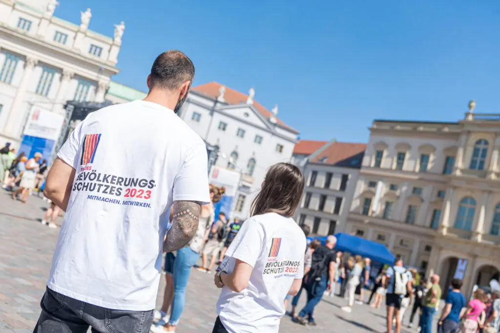 Zwei Personen mit weißen T-Shirts mit dem Aufdruck „Bevölkerungsschutz 2023 – Mitmachen. Mitwirken.“ stehen auf dem Platz vor dem Potsdamer Alten Markt, im Hintergrund viele Besucher*innen und historische Gebäude.