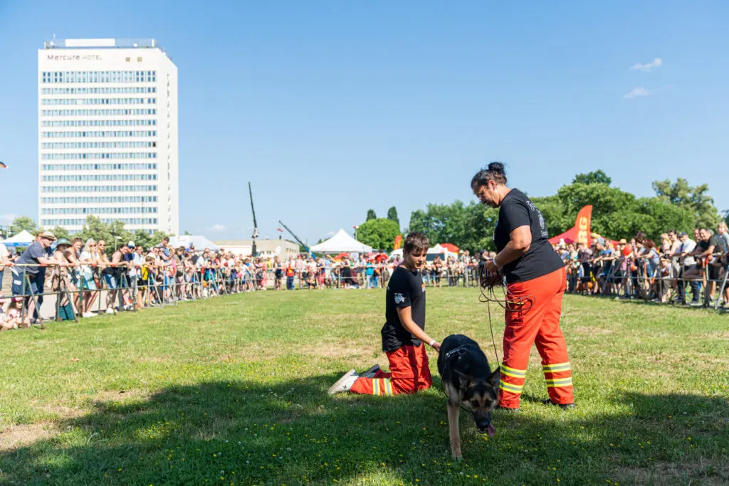 Zwei Rettungshundführer*innen demonstrieren die Arbeit mit einem ausgebildeten Suchhund auf einer Wiese, umringt von einem großen, interessierten Publikum. Im Hintergrund ist ein hohes Gebäude mit der Aufschrift "Mercure Hotel" zu sehen.