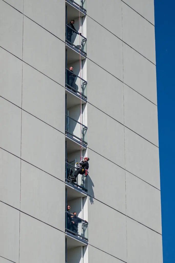 Höhenretter*in seilt sich an der Fassade eines Hochhauses ab, während mehrere Personen auf den angrenzenden Balkonen das Geschehen beobachten. Die Übung findet bei strahlend blauem Himmel statt.