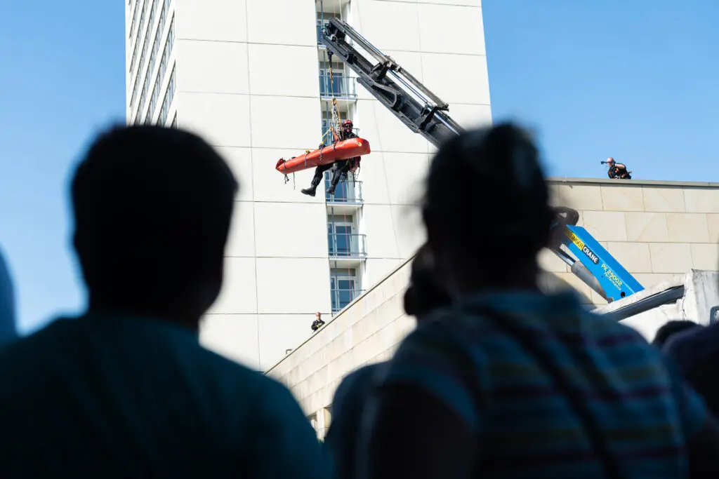 Zuschauer*innen beobachten eine Höhenrettungsübung an einem Hochhaus, bei der Einsatzkräfte eine Person in einer Trage mithilfe eines Krans abseilen.