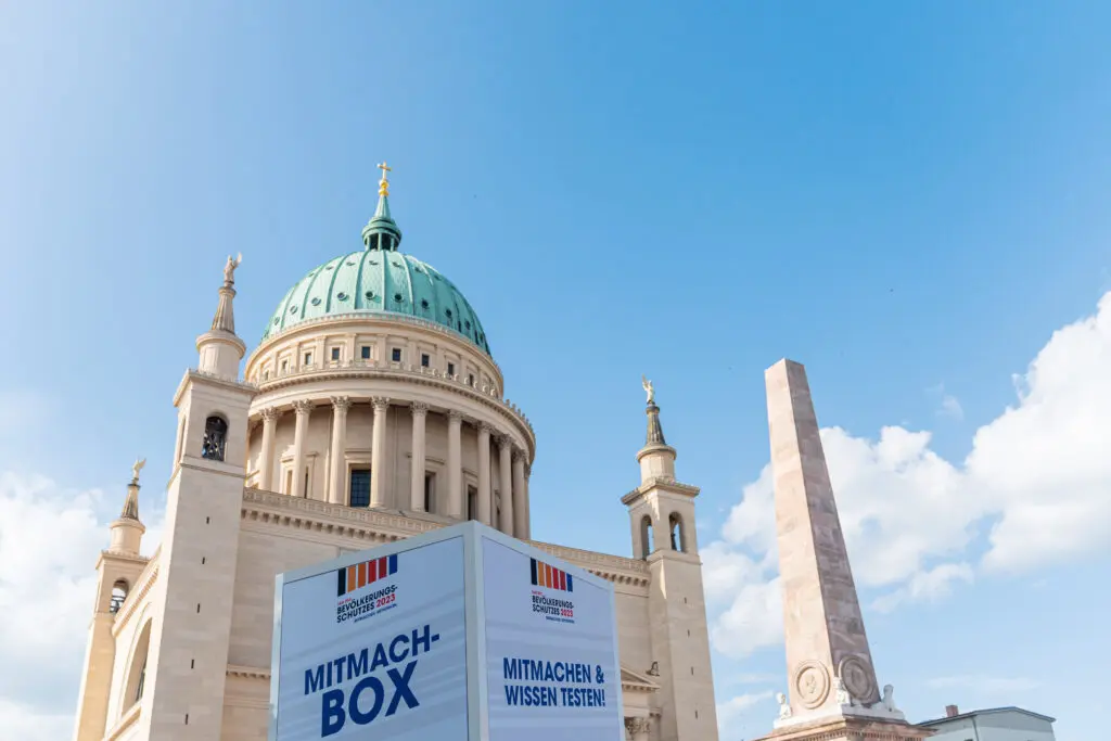 ChatGPT:
Die Mitmach-Box zum Bevölkerungsschutz 2023 steht vor der imposanten Kuppel der St.-Nikolaikirche in Potsdam, daneben ein hoher Obelisk – beide markante Wahrzeichen des Alten Marktes.