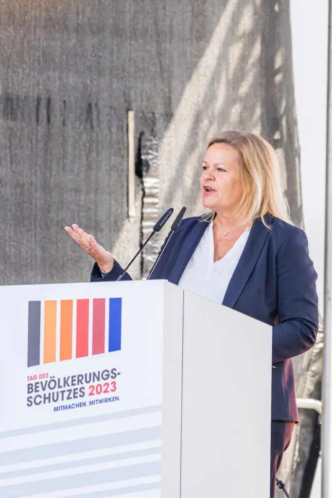 Bundesinnenministerin Nancy Faeser spricht auf einer Bühne am Tag des Bevölkerungsschutzes 2023 in Potsdam. Sie steht hinter einem Rednerpult mit Veranstaltungslogo und gestikuliert mit der linken Hand.