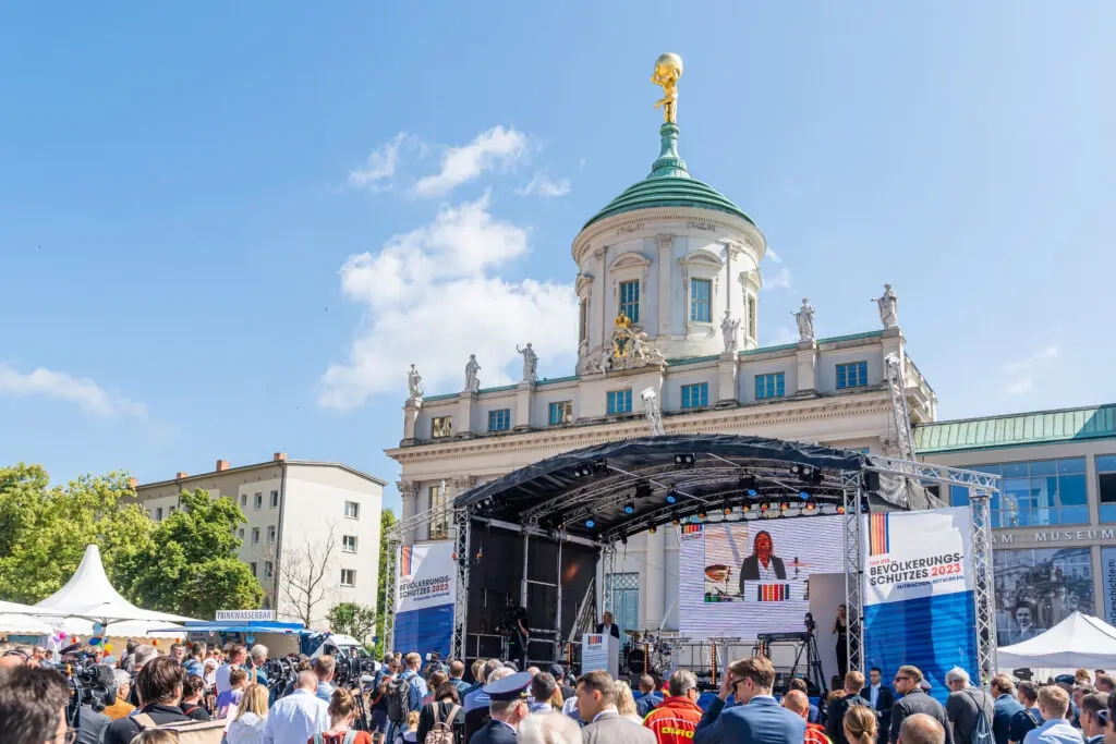 Blick auf eine große Bühne mit LED-Leinwand beim Tag des Bevölkerungsschutzes 2023 in Potsdam, vor dem markanten Gebäude mit goldener Kuppel. Viele Menschen stehen vor der Bühne und verfolgen das Geschehen bei strahlendem Sonnenschein.