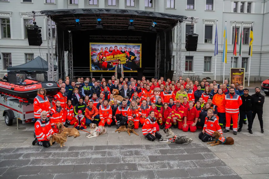 Gruppenbild zahlreicher Einsatzkräfte mit Rettungshunden vor einer Bühne beim Katastrophenschutztag, im Hintergrund ein farbenfrohes Banner mit dem Titel „Held*innen des Alltags“. Links steht ein Rettungsboot auf einem Anhänger.