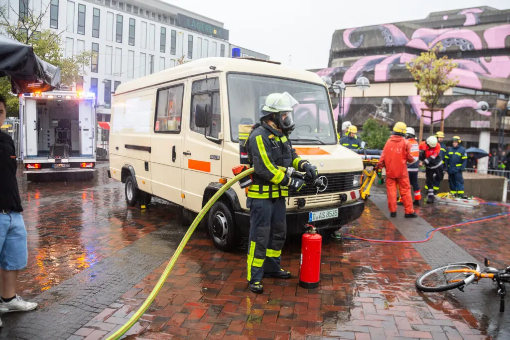 ChatGPT:
Ein Feuerwehrmann in voller Schutzausrüstung steht bei Regen vor einem alten weißen Einsatzfahrzeug und hält einen Feuerwehrschlauch sowie einen Feuerlöscher. Im Hintergrund arbeiten weitere Einsatzkräfte an einem Übungsszenario mit einem gestürzten Fahrrad, während ein Gebäude mit großflächiger Graffitikunst zu sehen ist.