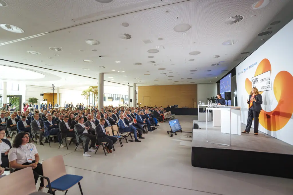 Großer Konferenzraum mit Publikum in Anzügen, Blickrichtung zur Bühne mit Rednerpult und Monitor.