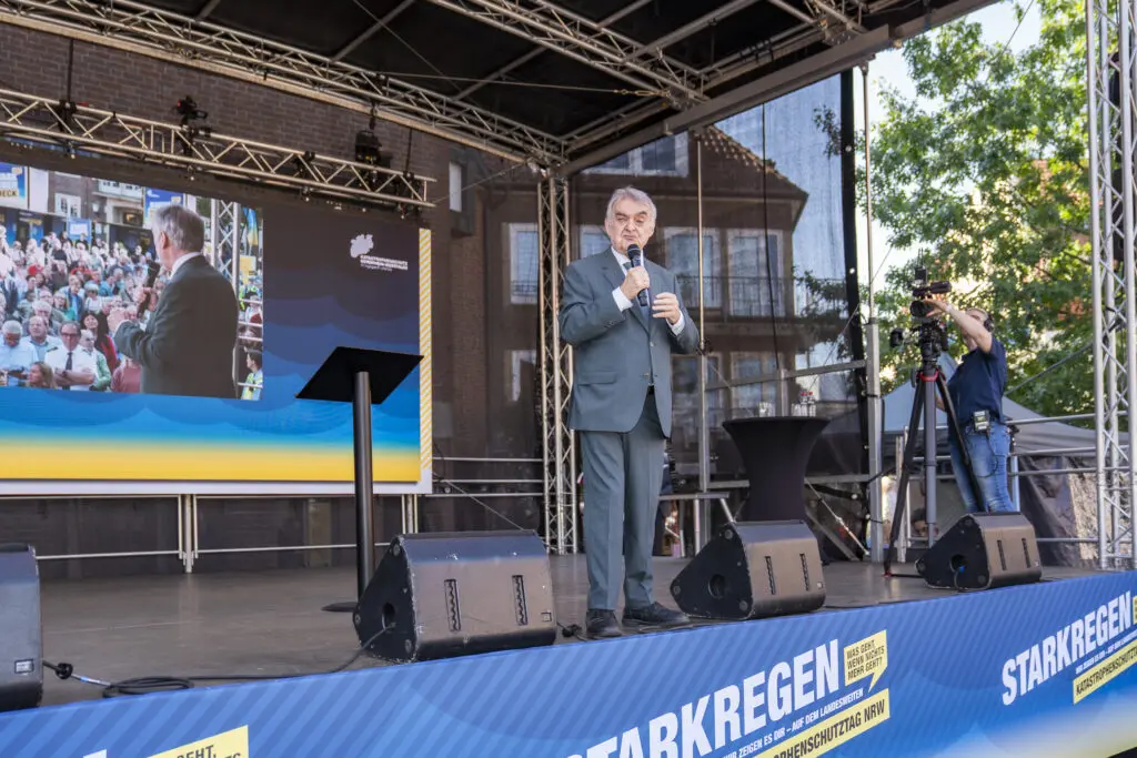 Ein Redner im Anzug spricht auf einer Bühne beim Katastrophenschutztag NRW in Coesfeld, im Hintergrund eine große LED-Leinwand mit seiner Live-Übertragung und dem Schriftzug „Starkregen“. Rechts steht ein Kameramann bei der Aufnahme.