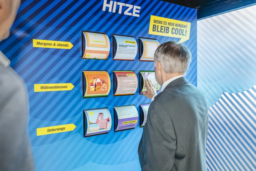 ChatGPT:
Ein Mann in Anzug steht vor einer interaktiven Wand mit dem Titel „Hitze – Wenn es heiß wird: Bleib cool!“. Die Wand enthält aufklappbare Infofelder mit Tipps für Hitzeschutz am Morgen, Mittag und unterwegs, bebildert mit anschaulichen Illustrationen.