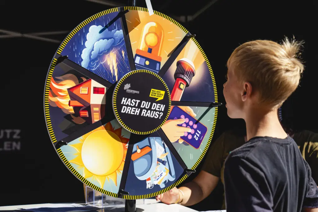 Ein Kind dreht an einem Glücksrad mit dem Slogan „Hast du den Dreh raus?“ – jede Sektion zeigt ein Symbol rund um Katastrophenschutz und Vorsorge.
