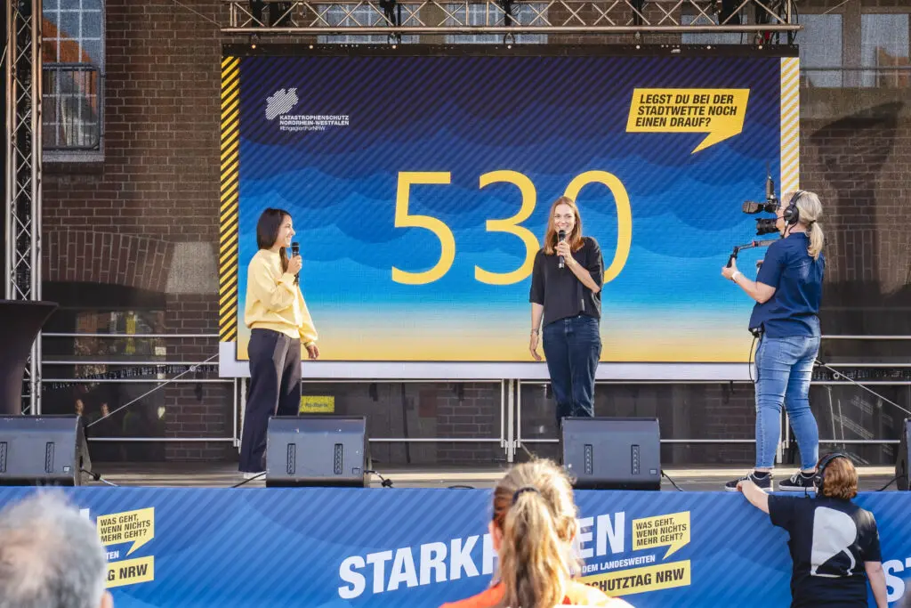Zwei Moderatorinnen stehen auf einer Bühne des Katastrophenschutztags NRW und präsentieren die Zahl „530“ auf einer großen LED-Leinwand, während eine Kamerafrau sie filmt – Teil der Aktion „Stadtwette“ mit dem Slogan „Legst du bei der Stadtwette noch einen drauf?“.