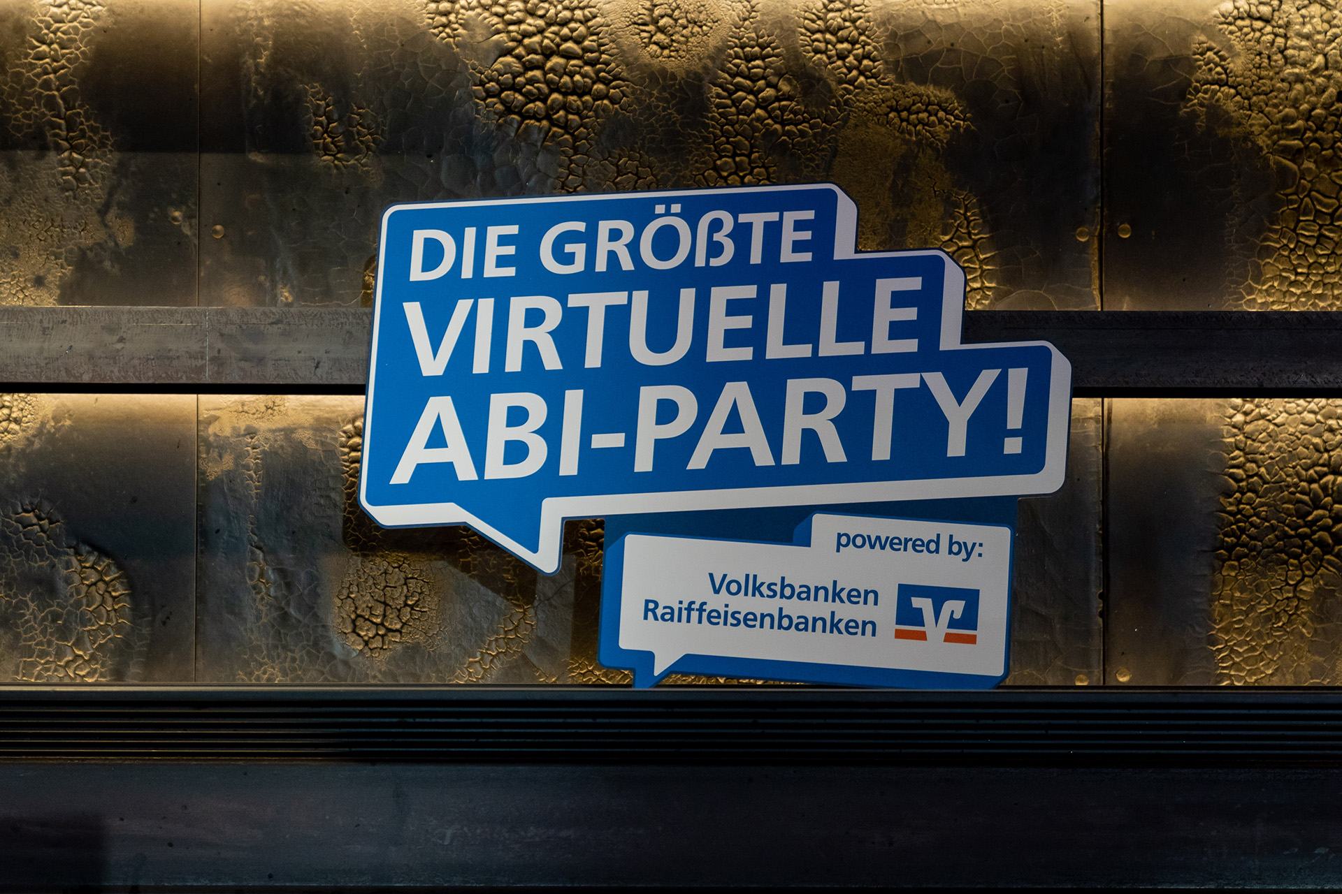 Schild mit Aufdruck „Die größte virtuelle Abi-Party!“ und Volksbanken Raiffeisenbanken-Logo vor strukturierter Wand.