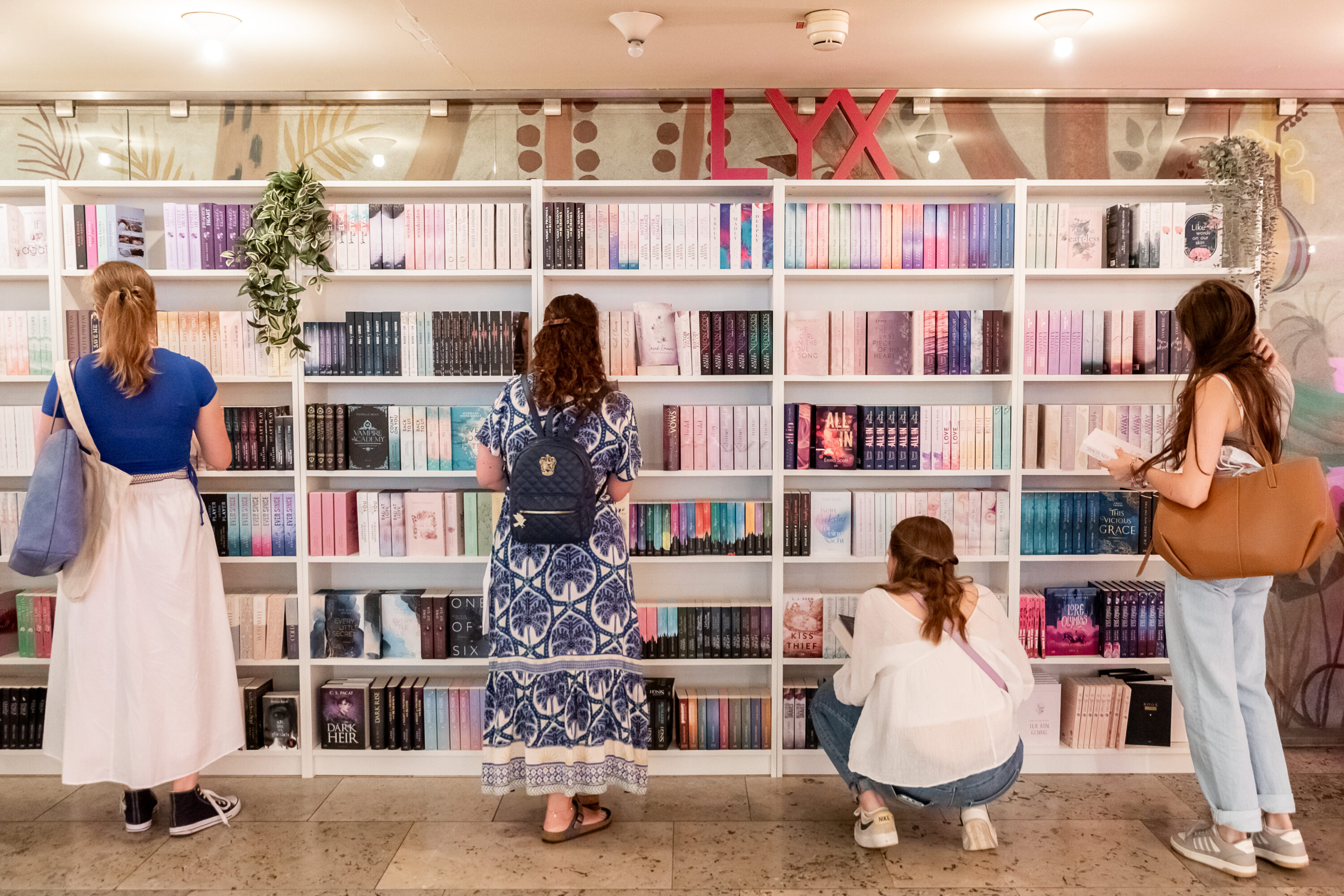 Fünf Frauen stöbern vor einem farbenfrohen Bücherregal mit LYX-Logo in einer hellen Eventlocation.