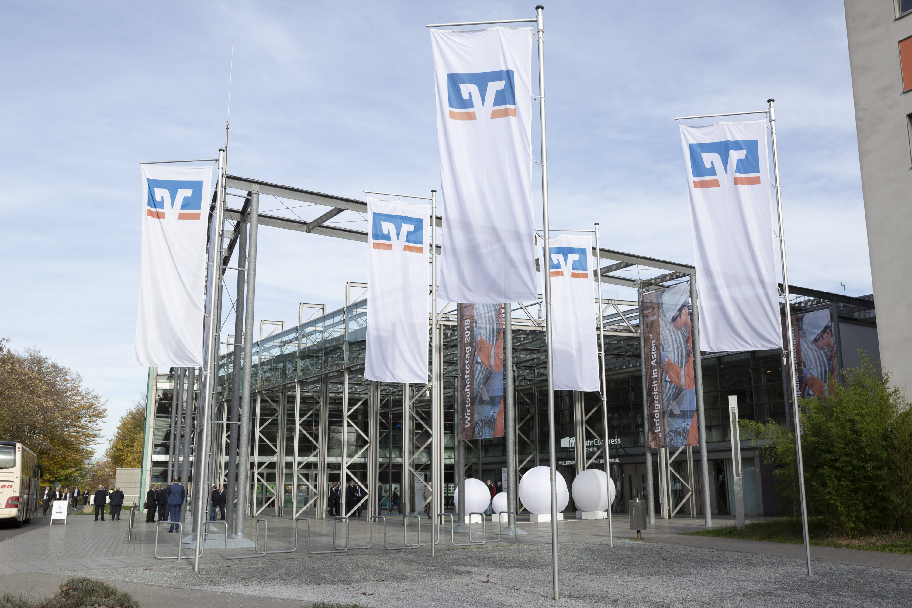 Außenaufnahme des Veranstaltungsorts mit modernem Glasbau, gesäumt von mehreren großen Volksbanken-Raiffeisenbanken-Fahnen. Einige Besucher*innen stehen vor dem Eingang, im Hintergrund ist ein Banner mit dem Titel „Erfolgreich in Asien“ zu sehen.