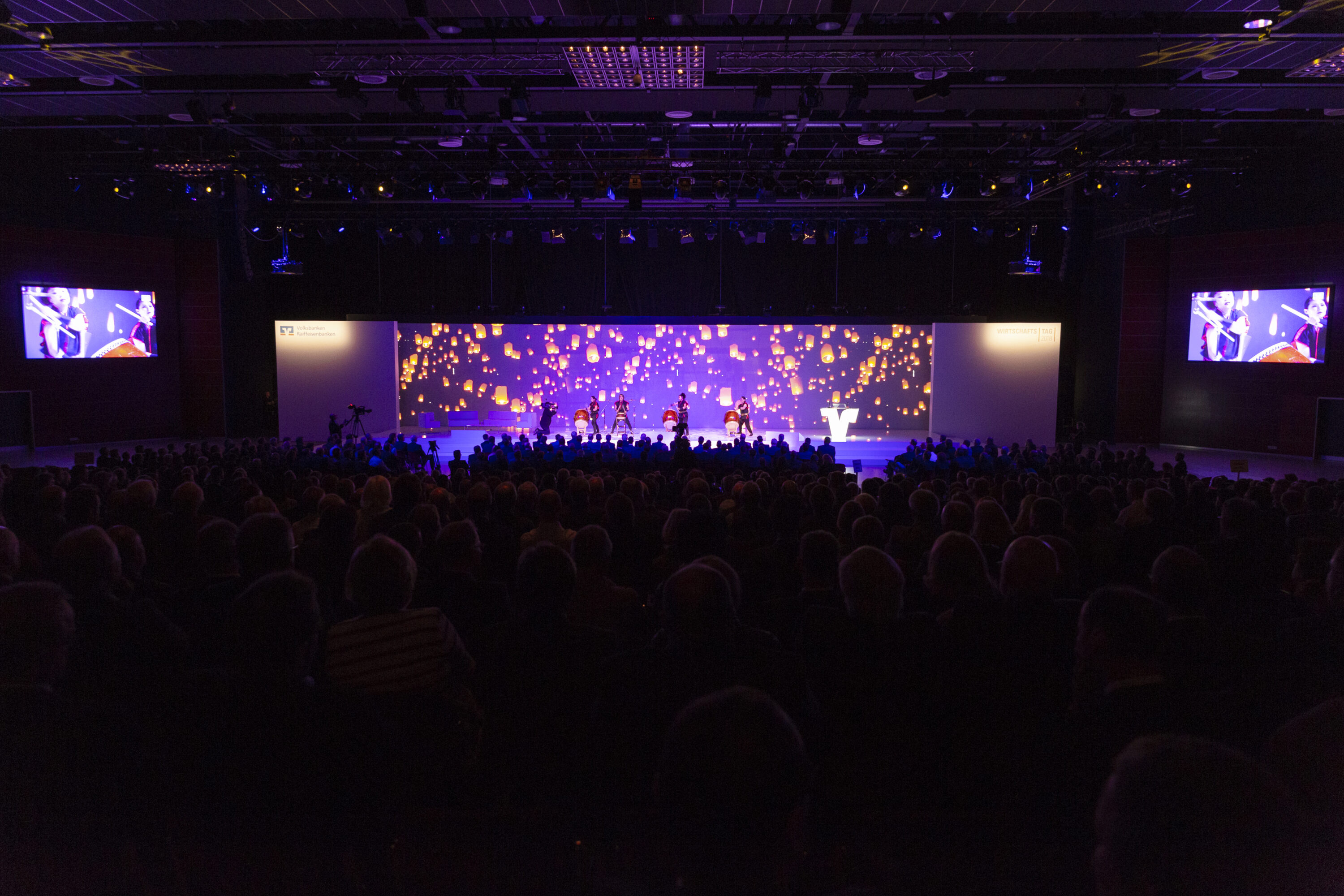 ChatGPT:
Großer Festsaal mit dunkler Bestuhlung, gefüllt mit Publikum, das einer hell erleuchteten Bühne mit Lichtshow und Tanzperformance zuschaut; auf den Seiten zwei Screens mit Nahaufnahmen der Künstler*innen.