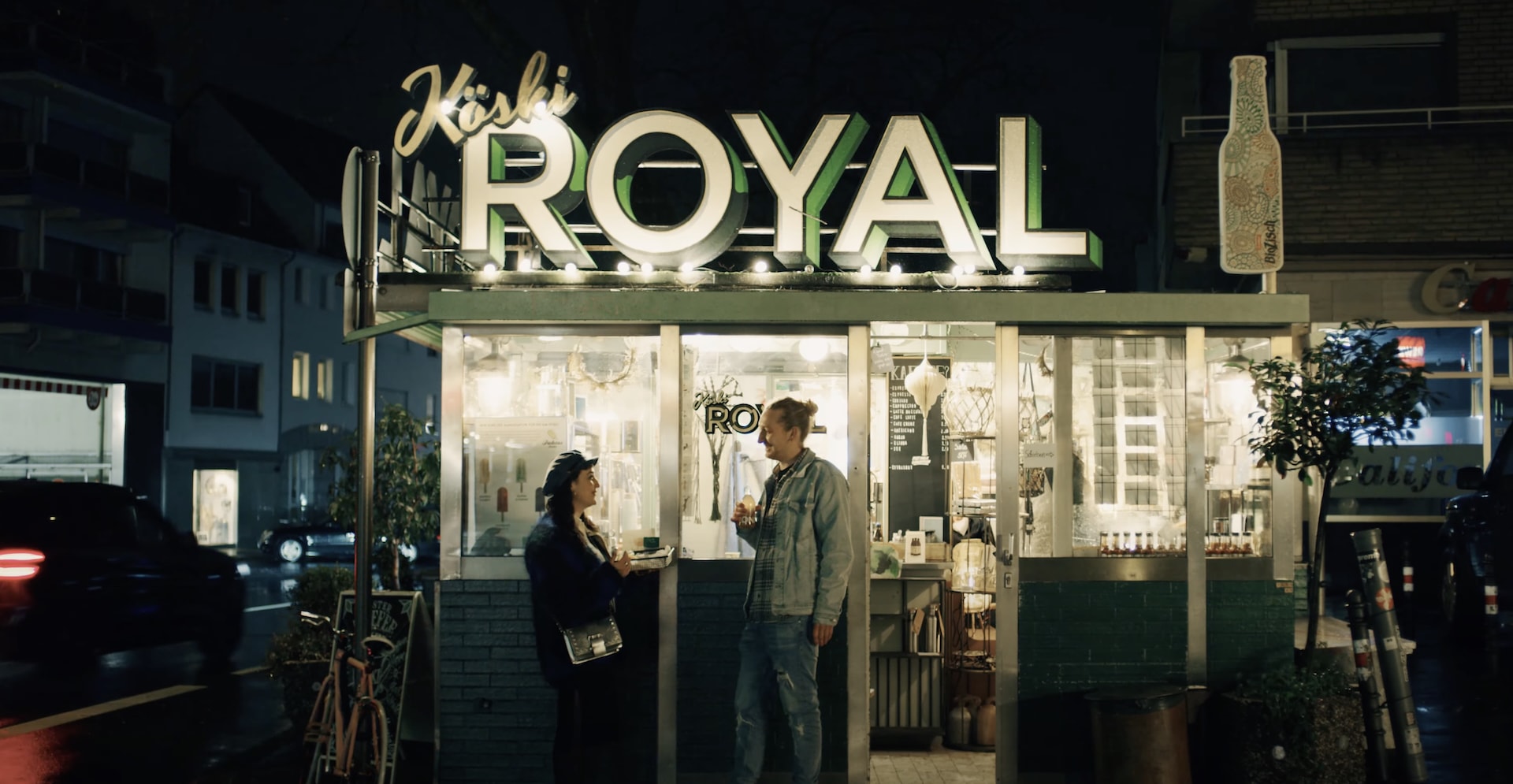 Nächtliche Szene vor einem Imbiss mit leuchtendem „Kölski Royal“-Schriftzug in Köln. Zwei Personen stehen davor, unterhalten sich und genießen Snacks und Drinks – das Setting wirkt urban und stimmungsvoll.