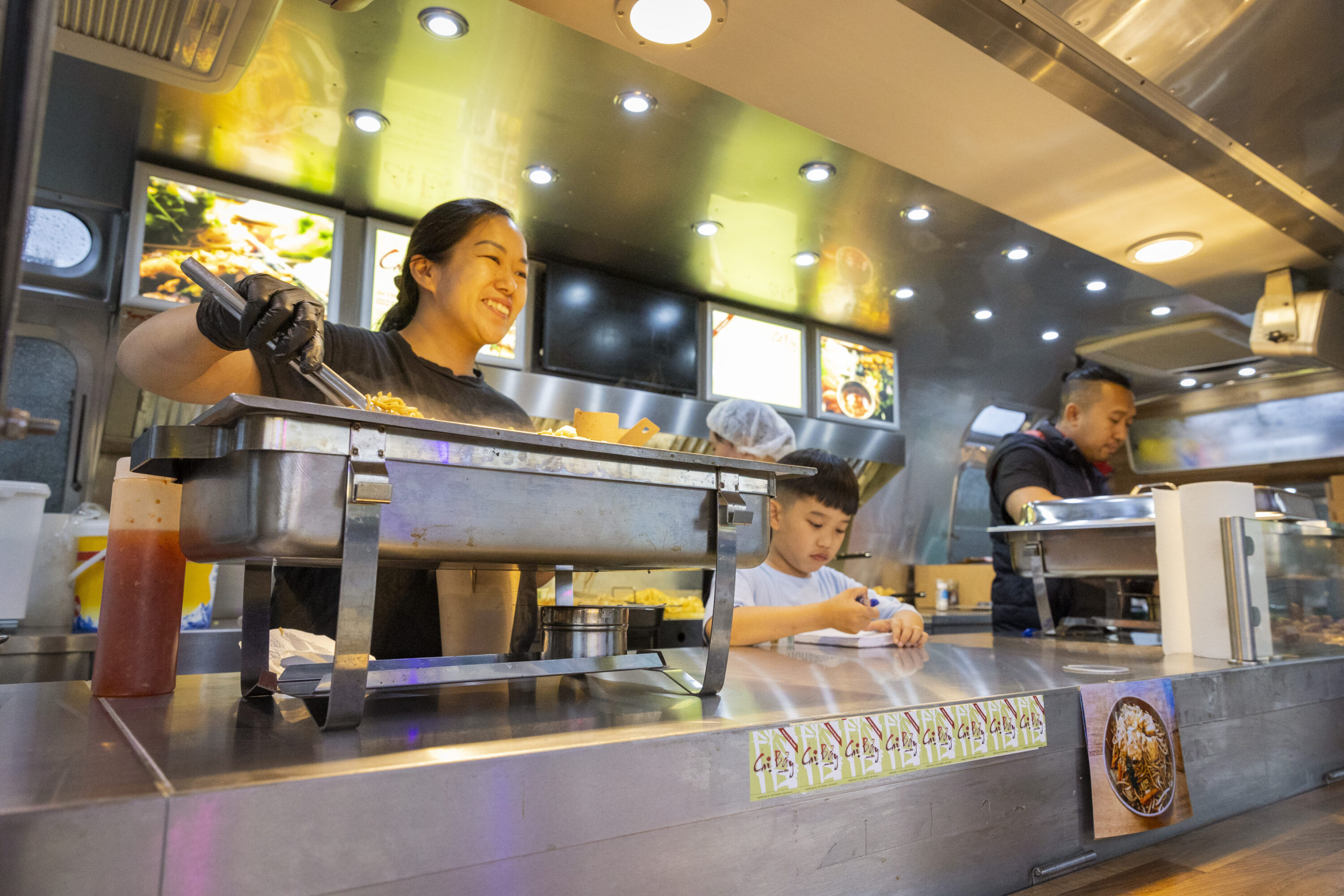 Drei Personen servieren asiatisches Streetfood aus einem Foodtruck, während dampfende Speisen im Vordergrund angerichtet werden.