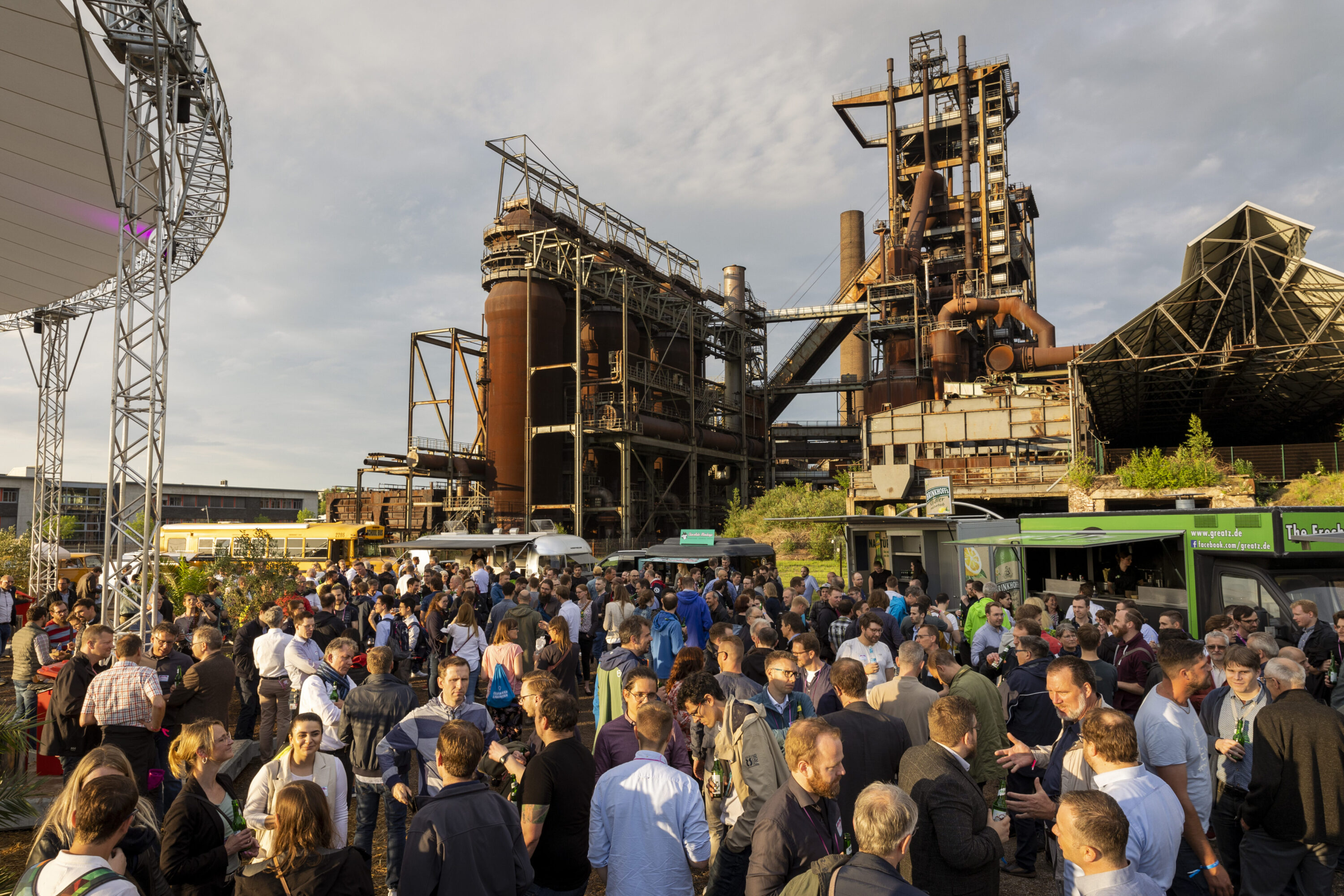 Große Menschenmenge bei einer Outdoor-Veranstaltung mit Foodtrucks vor einem imposanten alten Industriekomplex bei Tageslicht. Links im Bild ragt das weiße Zeltdach des Events ins Bild.