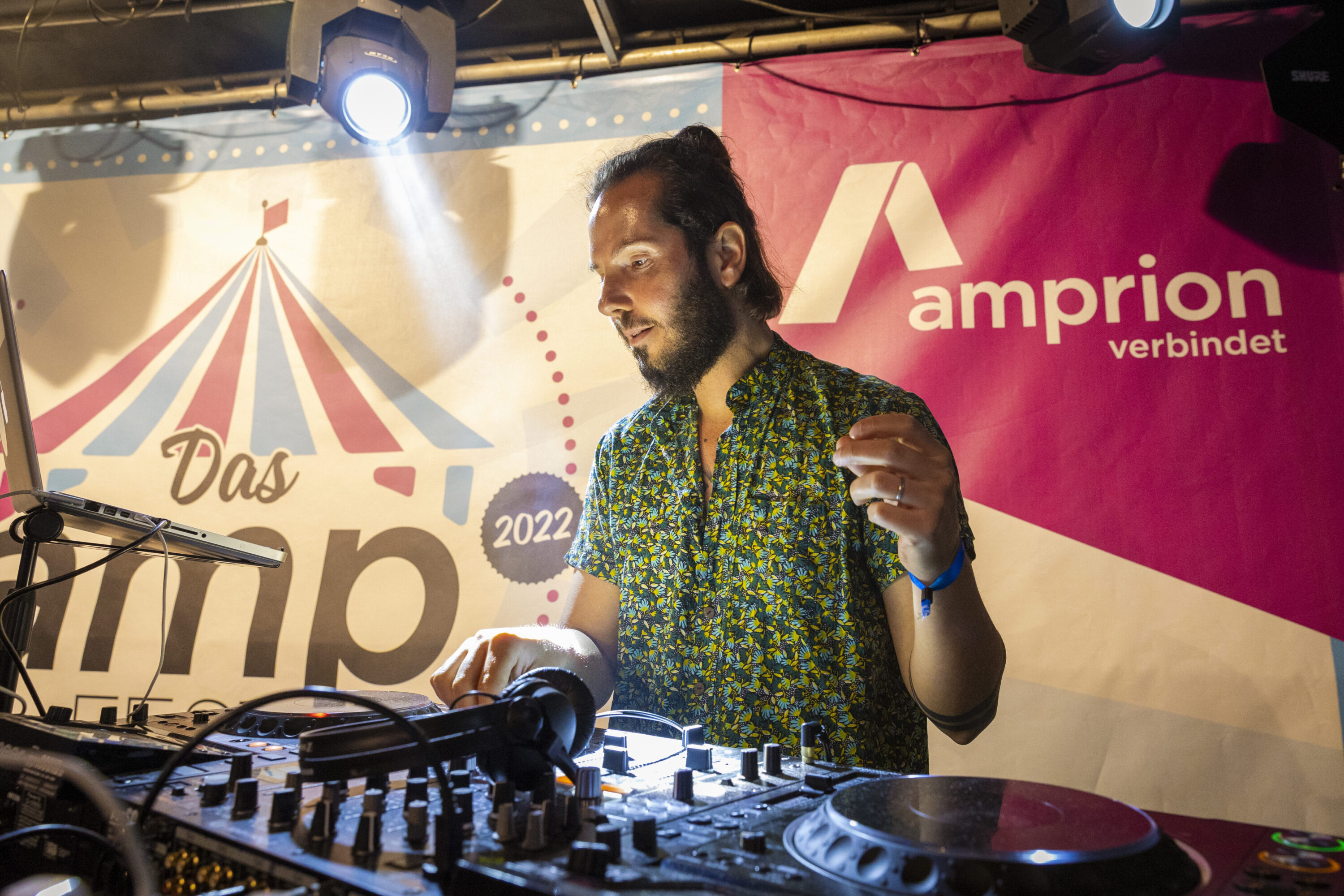DJ Mauro mit geblümtem Hemd steht konzentriert hinter seinem Mischpult auf dem amp Fest 2022, während bunte Scheinwerfer und ein Banner mit dem Amprion-Logo die Bühne beleuchten.