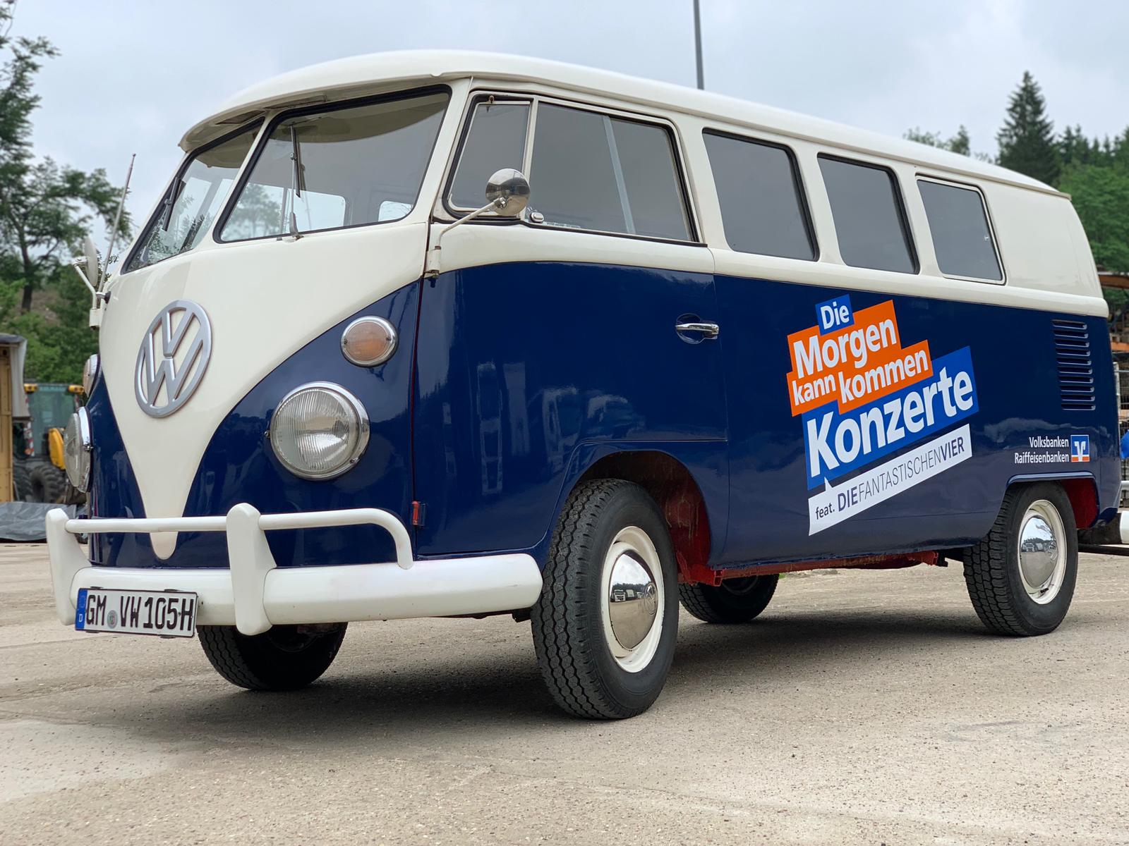 Ein alter blau-weißer VW-Bus mit einem deutschen Werbeschild auf der Seite, das für Konzerte mit den Volksbanken Raiffeisenbanken wirbt, ist im Freien auf einer Betonfläche geparkt.